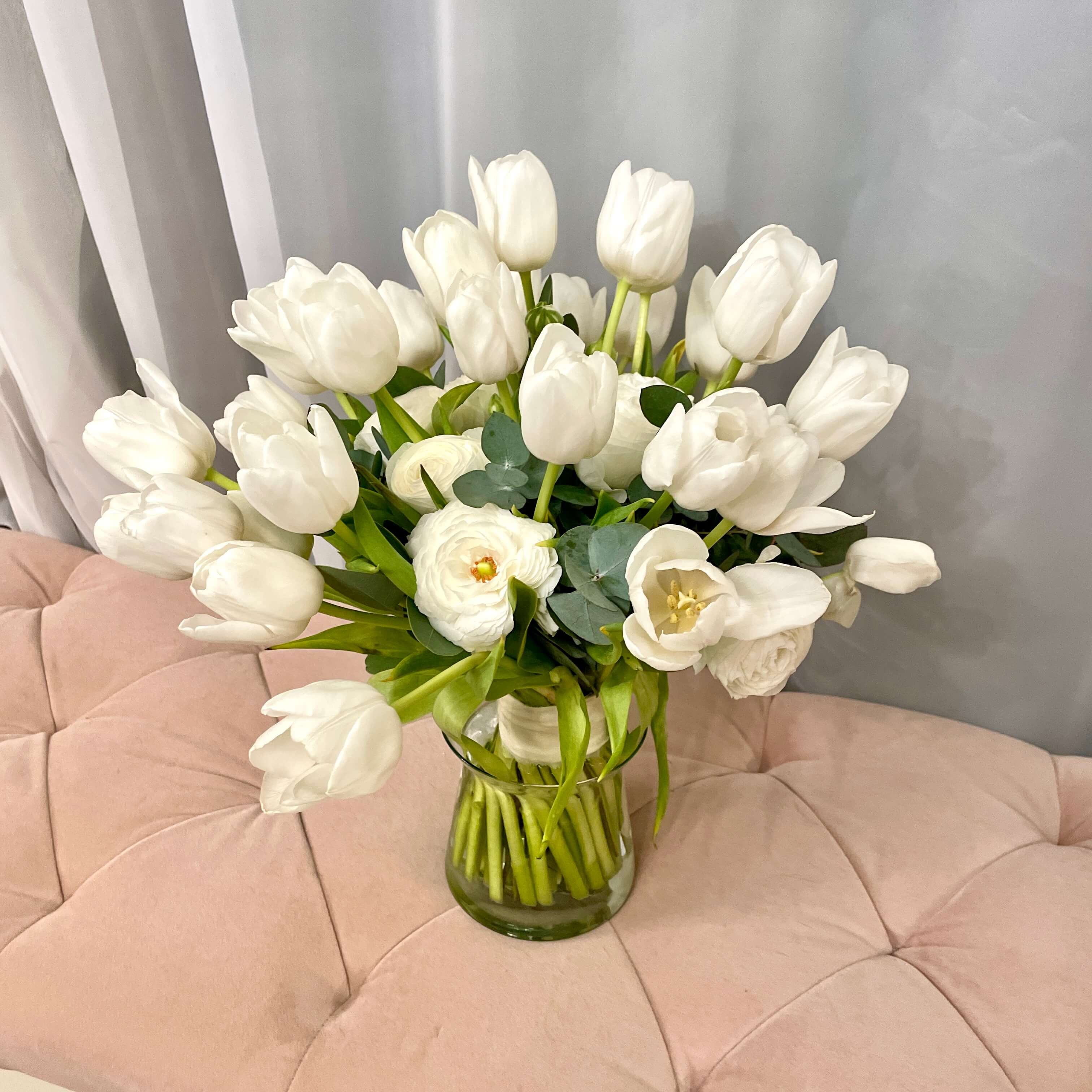 Bouquet of white tulips in a clear vase on a pink cushioned surface.