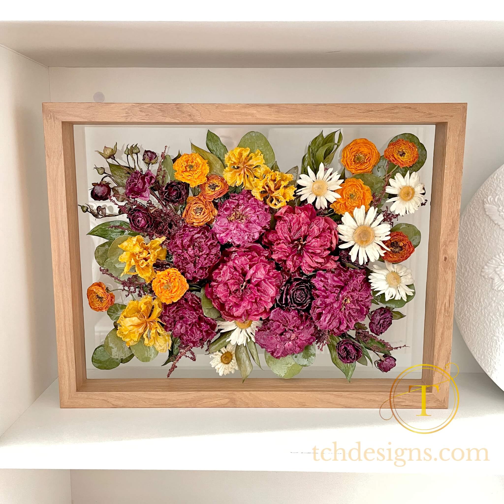 Framed arrangement of colorful flowers on a white shelf with 'tchdesigns.com' branding.