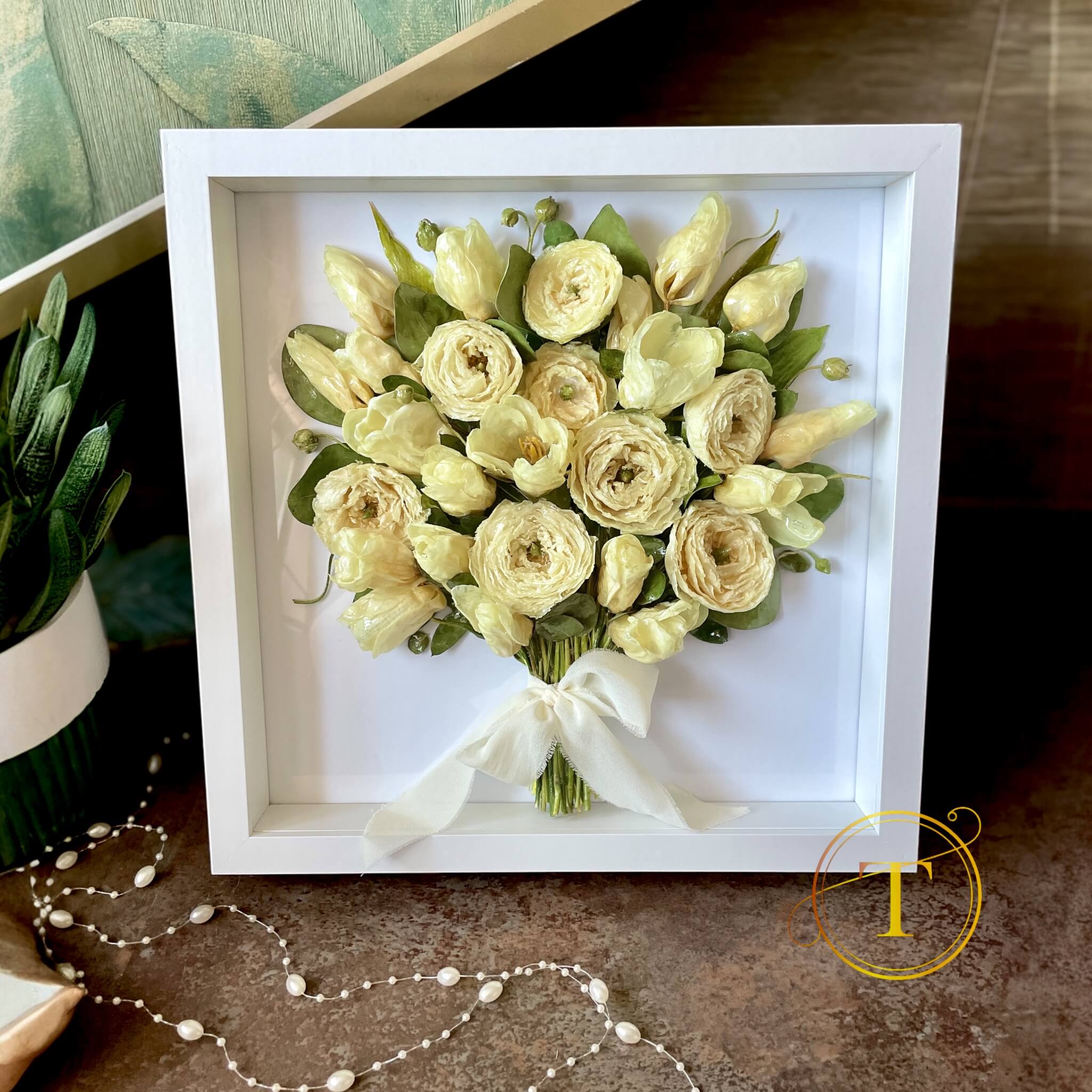 Framed bouquet of light-colored flowers with a white ribbon on a textured surface.