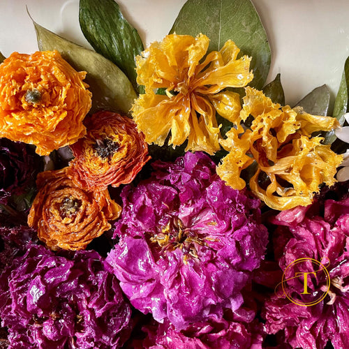Close-up of dried epoxy coated flowers in orange, yellow, and purple with green leaves on a white background.