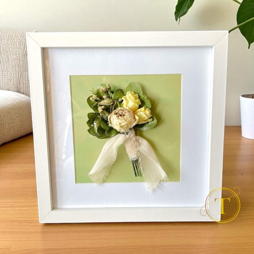 Framed floral corsage on a wooden surface with a plant in the background